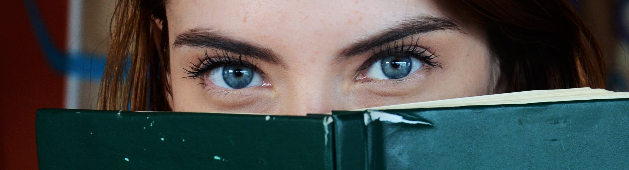 Close-up of a woman with blue eyes peeking over a book, portraying curiosity.